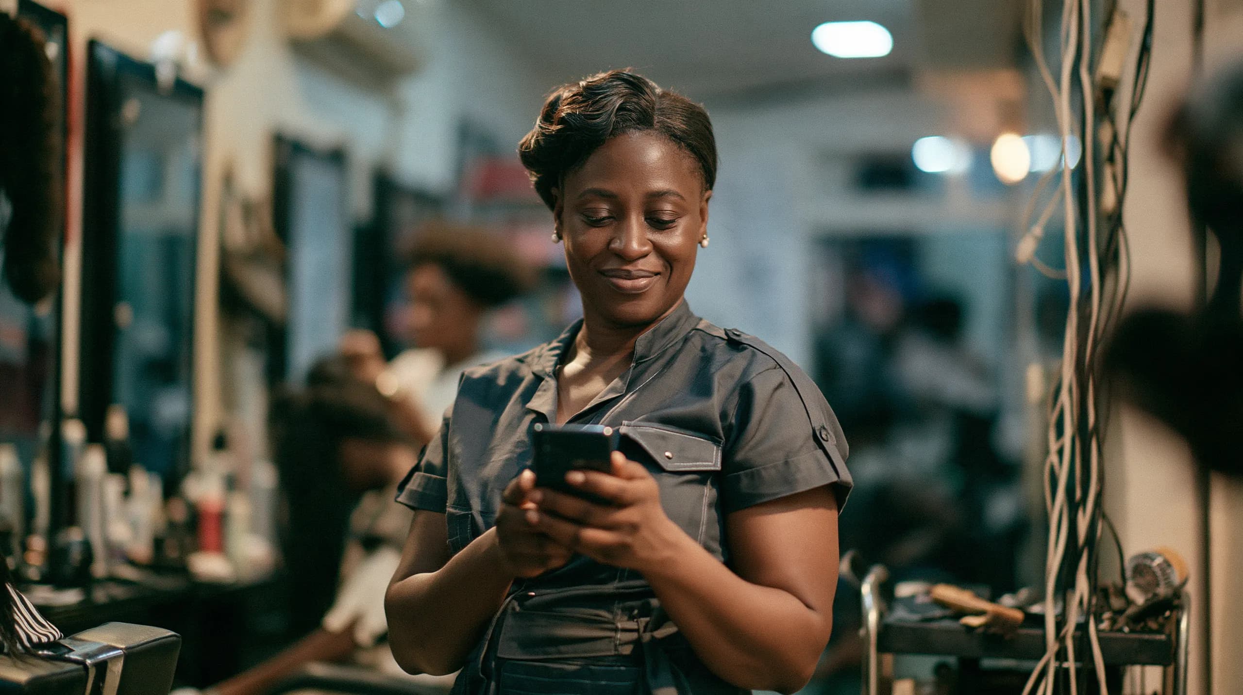 A salon owner checks her phone between clients, her shop busy behind her.