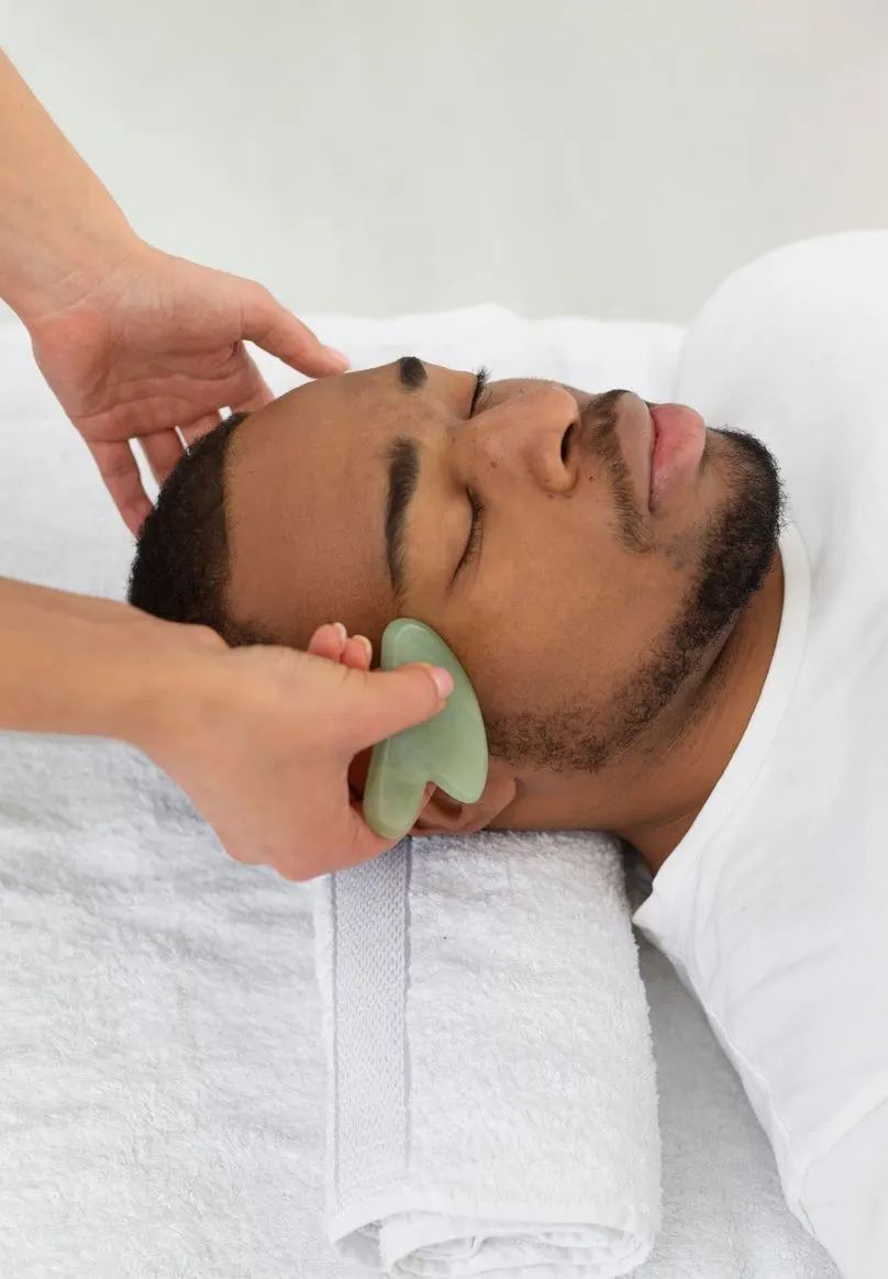 A therapist works a jade gua sha across a man's cheek during a facial.